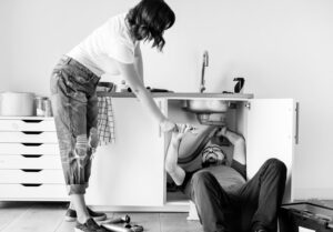 Man onder gootsteen voert reparatie uit terwijl vrouw hem gereedschap aanreikt tegen vochtproblemen