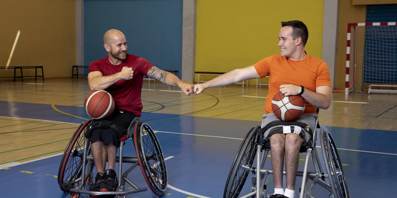 Twee mensen in sportrolstoelen met een basketbal in een sporthal.