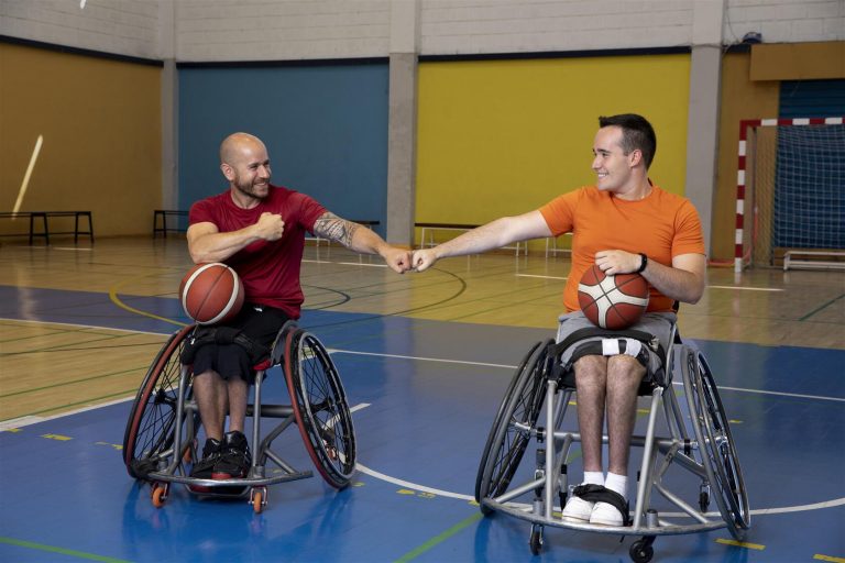 Twee mensen in sportrolstoelen met een basketbal in een sporthal.