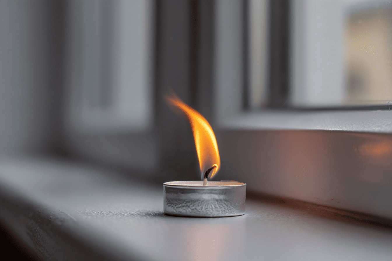 Brandend waxinelichtje bij een raamkozijn waarvan de vlam opzij buigt